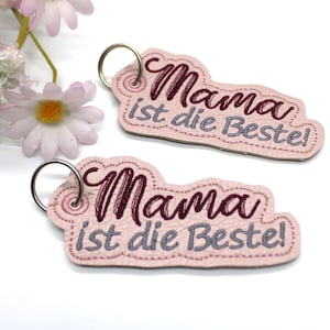 May include: Two pink keychains with the text "Mama ist die Beste!" embroidered in burgundy and gray. Each keychain has a silver-colored ring. The keychains are shaped like a rounded rectangle.