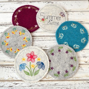 May include: Six embroidered felt coasters with various designs. One coaster is red with a white circle of flowers, one is white with a flower design, one is gray with a purple flower design, one is gray with a yellow flower design, one is blue with white butterflies, and one is white with the text "Summer Time" in black.