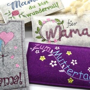 May include: Four felt pouches with embroidered designs for Mother's Day. The pouches are in gray, white, purple, and light gray. The designs include hearts, flowers, and the words "Mama", "For Mama", "Zum Muttertag", and "Mama du bist wundervoll!"