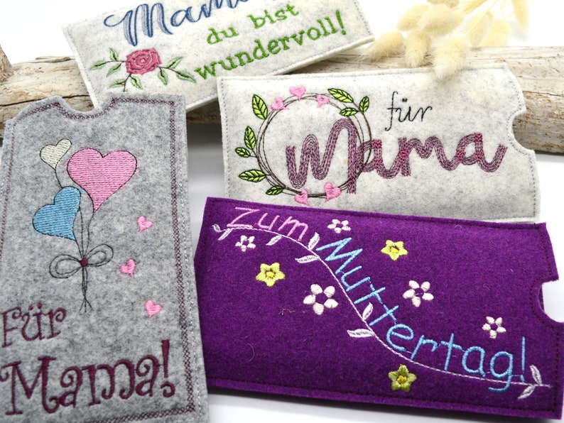 May include: Four felt pouches with embroidered designs for Mother's Day. The pouches are in gray, white, purple, and light gray. The designs include hearts, flowers, and the words "Mama", "For Mama", "Zum Muttertag", and "Mama du bist wundervoll!"