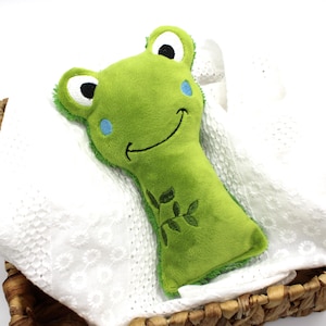 May include: A green plush frog toy with a smiling face, blue accents, and embroidered leaf details. The toy is resting on a white, textured surface inside a woven basket. The frog's eyes are black and white.