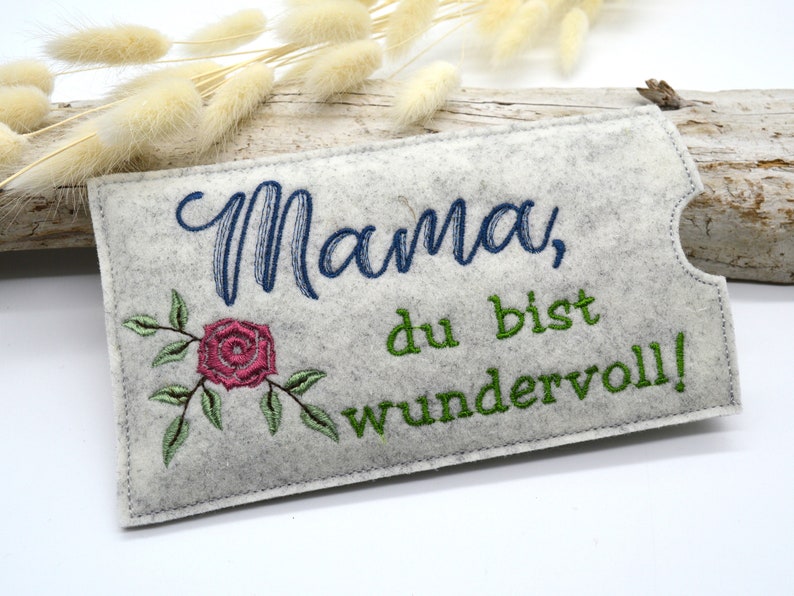 May include: A gray felt pouch with a white embroidered rose and the text "Mama, du bist wundervoll!" in green and blue thread.