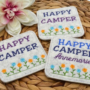 以下が含まれることがあります： 「Happy Camper」と刺繍されたグレーのフェルトコースター3枚。右側のコースターには、テキストの下に「Annemarie」という名前が刺繍されています。3つのコースターすべてに、青、オレンジ、黄色の刺繍された花が並んでいます。