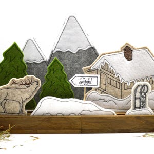 May include: Felt Christmas scene with a brown cabin, two green trees, a white mountain range, a brown deer, and a sign that says "Gipfel".