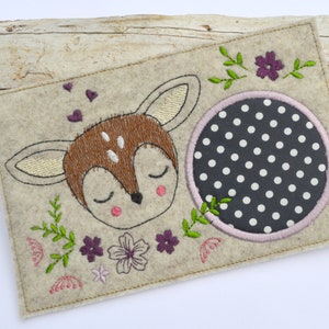 May include: A beige felt applique with a brown and white deer with closed eyes, a black and white polka dot circle, and green leaves and purple flowers.