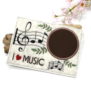 May include: A cream-colored felt music-themed item with a brown circular opening. It features a treble clef, musical notes, and the text "I ❤️ MUSIC" in black and red. Green leaf accents surround the circular opening and musical notes.