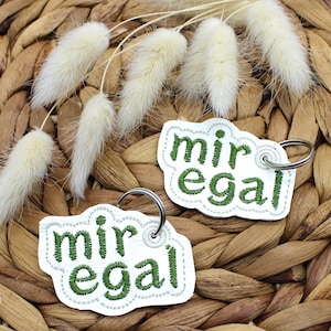 May include: Two white keychains with the German phrase "mir egal" embroidered in green. The keychains have a silver ring and a decorative border. The background features dried white flowers and a woven basket.