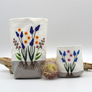 Può includere: Due contenitori in tessuto bianco con ricami floreali. Il contenitore più grande presenta fiori blu, arancioni e viola con steli verdi e radici marroni. Il più piccolo ha fiori blu e rosa. Entrambi hanno una base grigia.