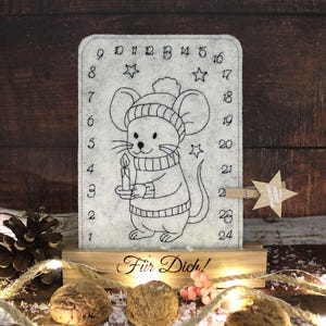 Könnte beinhalten: Grauer Adventskalender aus Filz mit einer handgezeichneten Maus, die eine Kerze hält. Der Kalender hat die Zahlen 1-24, Sterne und den Text "Für Dich!" auf dem Holzsockel. Ein sternförmiges Etikett mit "Merry Christmas" ist angebracht.