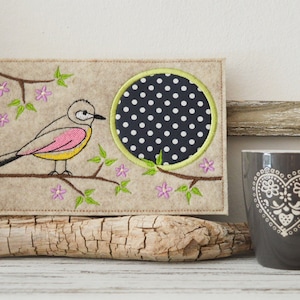 Peut inclure: Décoration murale en feutre brodée avec un oiseau perché sur une branche avec des fleurs roses. L'arrière-plan est un cercle à pois noir et blanc.