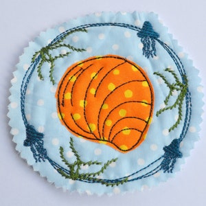 Può includere: Un cerotto rotondo in tessuto blu con pois bianchi, che presenta una conchiglia arancione e gialla con cuciture nere e alghe verdi intorno ai bordi.