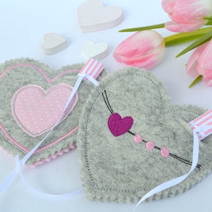 May include: Two gray felt heart-shaped pouches with pink and white ribbon ties. One pouch has a pink heart with white polka dots and the other has a pink heart with a black outline and pink dots.