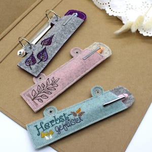 May include: Three felt pen holders in grey, pink, and teal, each with an embroidered design. The grey holder features purple leaves, the pink one has a branch, and the teal one has text and mushrooms. Each has a metal clip.