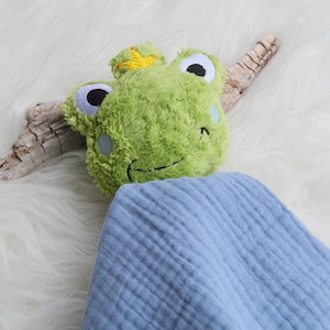 May include: A green plush frog with a yellow crown and blue eyes is attached to a blue muslin baby blanket.
