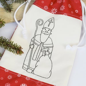 Pode incluir: Uma sacola de presente de tecido branco com um padrão de floco de neve vermelho e branco na parte inferior. A sacola apresenta uma imagem bordada em preto e branco de São Nicolau segurando um saco e um cajado.