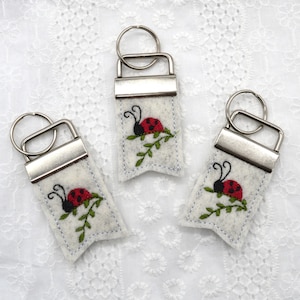 May include: Three white felt keyrings with silver key rings. Each keychain features a red and black ladybug embroidered on a green stem with leaves.