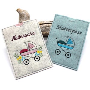Könnte beinhalten: Zwei Filz-Mutterpasshüllen. Eine ist hellgrau mit einem rosa und gelb geblümten gestickten Kinderwagendesign. Die andere ist hellblau mit einem blauen gestickten Kinderwagendesign. Das Wort "Mutterpass" ist auf beiden gestickt.