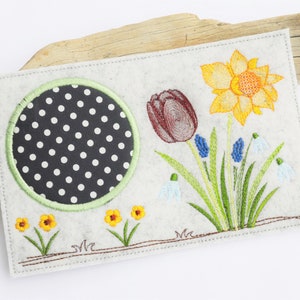 May include: A white felt card with embroidered spring flowers. The card features a black and white polka dot circle, a red tulip, a yellow daffodil, blue grape hyacinths, and white snowdrops.