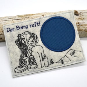 Peut inclure: Un patch en feutre blanc avec une broderie noire d'un sac à dos, de chaussures de randonnée et d'une scène de montagne. Le texte "Der Berg ruft!" est brodé en violet sur le patch. Un cercle en feutre bleu se trouve dans le coin supérieur droit du patch.