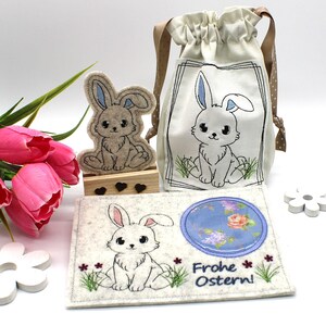 May include: An Easter-themed collection featuring a felt bunny figurine, a drawstring bag with a bunny illustration, and a felt card with a bunny and the text "Frohe Ostern!" The items are in neutral tones with blue and pink accents.