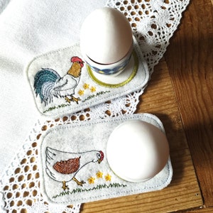 May include: Two felt egg holders with embroidered designs. One holder features a rooster with a red comb and a green tail. The other holder features a brown hen. Both holders have a white egg inside.