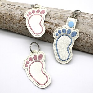 May include: Two white leather keychains with embroidered baby footprints. One keychain has a pink footprint and the other has a blue footprint.