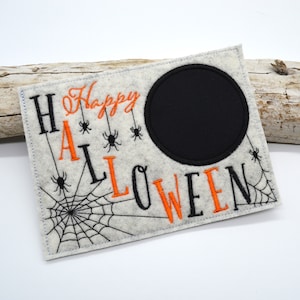 Peut inclure: Une pancarte en feutre gris pour Halloween avec les mots "Happy Halloween" brodés en fil noir et orange. La pancarte a un cercle noir au centre et un motif de toile d'araignée en bas à gauche.
