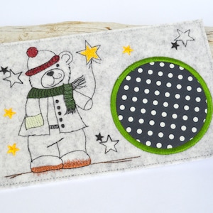 May include: A white felt card with a black and white polka dot circle in the center. The circle is surrounded by a green border. A white bear with a red hat and green scarf is holding a star on the left side of the card.