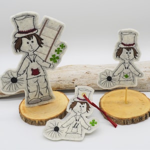 Może przedstawiać: Dwie filcowe figurki kominiarzy, jedna większa od drugiej, obie w czarnych kapeluszach i trzymające miotłę. Figurki stoją na drewnianych podstawach.