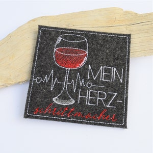 May include: Embroidered felt coaster featuring a red wine glass and a white EKG line with the text "Mein Herz-schrittmacher" in white.