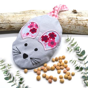 Può includere: Un giocattolo di stoffa grigio a forma di topo con orecchie a fiori rosa e un fiocco rosa e bianco a quadri. Il giocattolo è appoggiato su una superficie di legno con un mucchio di piccoli oggetti rotondi di colore marrone chiaro.