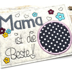 Könnte beinhalten: Ein weißes Filz-Schild mit dem Text "Mama ist die Beste!" in schwarzer Stickerei. Das Schild ist mit bunten gestickten Blumen und Herzen verziert. In der Mitte des Schildes befindet sich ein schwarz-weißer Kreis mit weißen Punkten und einem rosa Rand.