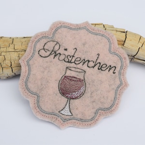 Może przedstawiać: Podkładka pod kubek z filcu w kolorze różowym z szarym haftowanym obramowaniem. Podkładka przedstawia kieliszek wina i napis "Prostchen".