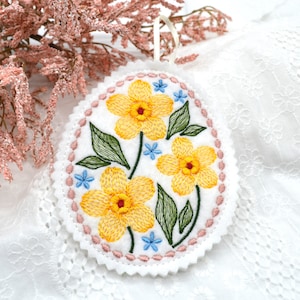 Pode incluir: Um ornamento oval de feltro branco com uma borda rosa festoneada. O ornamento é bordado com flores amarelas e azuis.