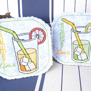 May include: Two embroidered coasters with a light blue background and scalloped edges. Each coaster features a glass of lemonade with ice cubes, a straw, and a citrus slice. The words "summer time" are embroidered vertically.