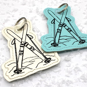 May include: Two ski-themed keyrings. One is off-white, the other is light blue. Each features a black embroidered design of crossed skis and poles in the snow. Both have a silver key ring attached at the top.