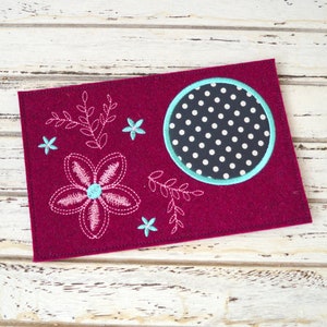 Peut inclure: Un objet rectangulaire en feutre bordeaux avec des motifs floraux brodés. Une grande fleur et plusieurs petites fleurs sont brodées en blanc et bleu clair. Un cercle avec un motif à pois est à droite.