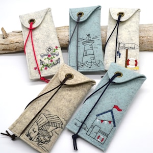 May include: Five felt eyeglass cases with different nautical embroidery designs. The cases are beige and light blue with a drawstring closure. The designs include an anchor, a lighthouse, a beach house, a sign that says "Strand", and a life preserver.