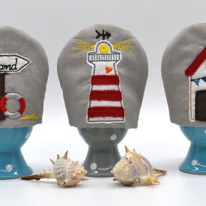 May include: Three gray egg cozies with embroidered nautical designs. The first cozie has a sign that says "Strand" with a life preserver and two fish. The second cozie has a red and white striped lighthouse with a yellow sun and a heart. The third cozie has a red and white beach house with a flag and bunting.