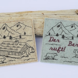 Puede incluir: Dos posavasos de fieltro con escenas de montaña bordadas. El primer posavasos es beige y presenta una cabaña con una cordillera de montañas en el fondo. El segundo posavasos es azul claro y presenta una cordillera de montañas con el texto "Der Berg ruft!" en marrón.