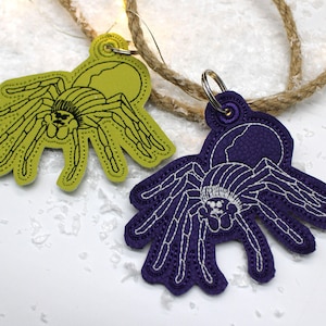 May include: Two embroidered spider keychains. One is lime green with black stitching, the other is purple with white stitching. Both have a silver key ring attached. The keychains are on a white surface with a rope and fairy lights.