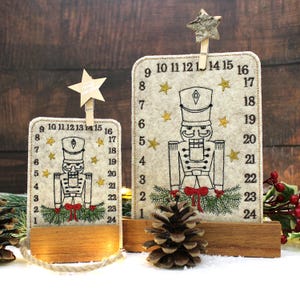 Könnte beinhalten: Zwei Adventskalender mit Holzsockel. Jeder Kalender hat eine Filztafel mit Nussknacker-Design, Zahlen und Sternen. Ein sternförmiger Clip hält die Oberseite jedes Kalenders. Der Text "Merry Christmas" ist auf dem Sternclip sichtbar.