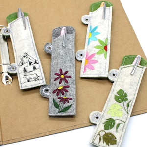 Könnte beinhalten: Eine Sammlung von Filz-Stifthaltern in verschiedenen Farben und Designs. Ein Halter ist grau mit gestickten Blumen, andere zeigen Berg- und Blumenmuster. Jeder Halter hat einen Stift und einen Metallring.