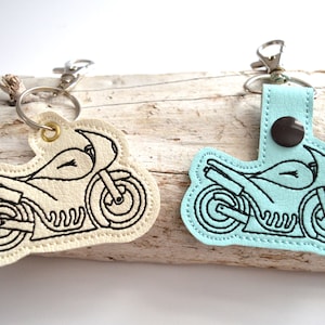 May include: Two motorcycle keychains, one in beige and one in light blue, with black embroidered outlines of a motorcycle.