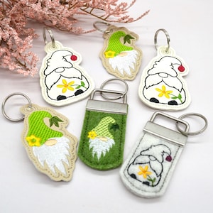 May include: Five different gnome-themed keychains. Each keychain features a gnome with a white beard and a red hat. The gnomes are wearing different colored hats and have different designs on their hats. The keychains are made of felt and are attached to silver key rings.