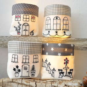 Könnte beinhalten: Vier dekorative Stoff-Kerzenhalter mit gestickten Haus- und Gartendesigns. Jeder Halter hat einen grau gemusterten oberen Teil und eine weiße Basis mit schwarzer Stickerei. Die Designs umfassen Fenster, Türen, Pflanzen und einen Tisch mit Stühlen.