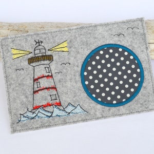 Könnte beinhalten: Eine graue Filzapplikation mit einem weißen und roten Leuchtturm mit gelben Lichtstrahlen und einem blauen Kreis mit weißen Punkten.