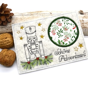 May include: A white felt card with a black and white embroidered nutcracker, a green circle with a white background and red berries, and the text "Schöne Adventszeit".