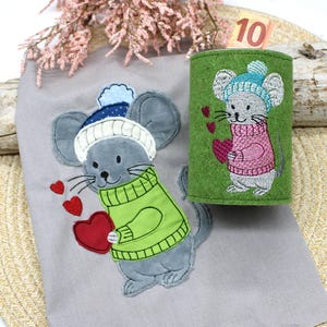 Könnte beinhalten: Ein gesticktes graues Mäuschen mit blau-weißem Hut und grünem Pullover, das ein rotes Herz hält, auf grauem Stoff. Ein grüner Filzzylinder mit Mäusemotiv und ein Zehn-Euro-Schein sind ebenfalls zu sehen. Thema: Valentinstag.