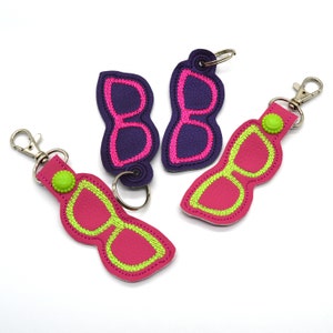 May include: Three keychains with a sunglasses design. One keychain is purple with pink stitching, and the other two are pink with green stitching.
